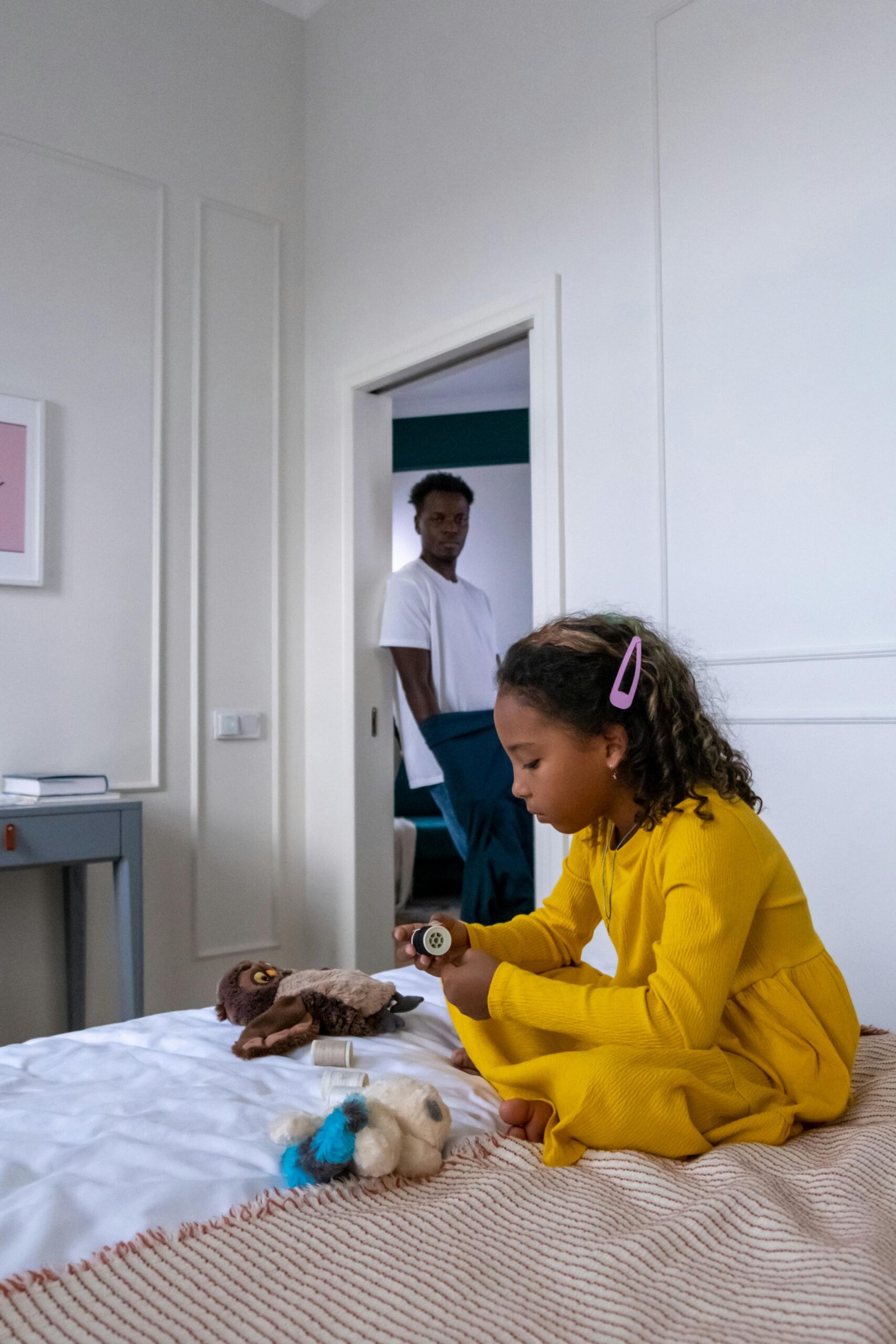 Young girl plays with toys on bed while father watches from the doorway.