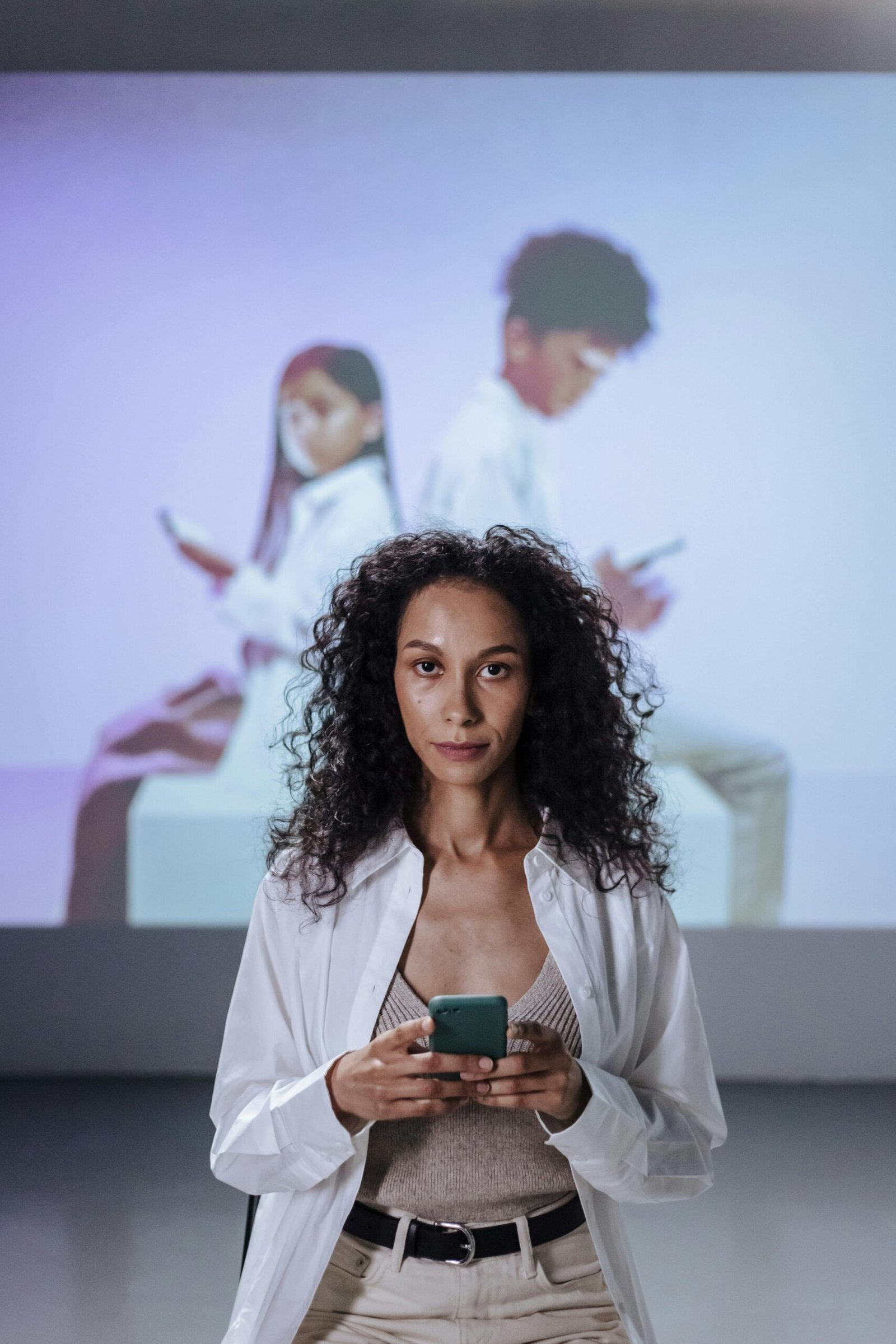 Confident woman holding smartphone with children projected on wall behind her, indoors.