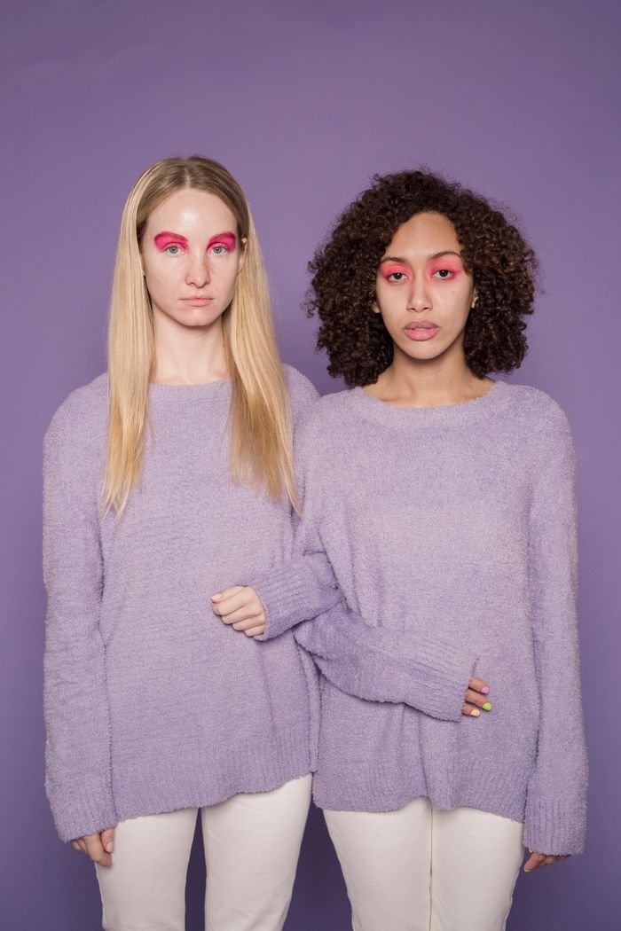 Multiracial women with pink eyeshadows in soft pullovers looking at camera in studio on purple background